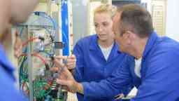 一名年轻的女工人和一名穿着蓝色工作服的年长的男工人在看一个工厂的电气开关板。