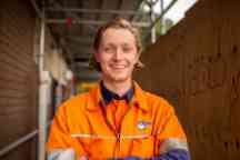 一个穿着橙色醒目工作衬衫的微笑的年轻人站在一个建筑工地前