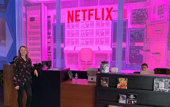 苏穿着黑衬衫和裤子,站在前面的Netflix接待区域。可以看到一个巨大的桌椅安装玻璃墙后面,空间被粉红色霓虹灯。