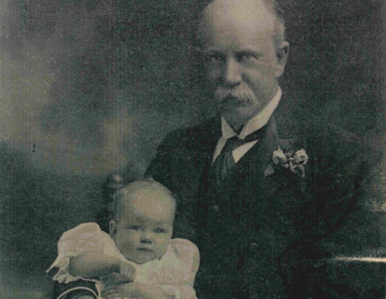 这是一张20世纪初乔治·史文朋勋爵和他的小女儿玛格丽特的照片，照片上的玛格丽特大约6个月大。她穿着一件白色褶边连衣裙坐在他的腿上，他的一只胳膊搂着她。