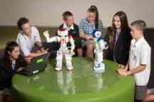 三个穿着校服的女孩和三个男孩围坐在一张绿色的桌子旁，和两个机器人玩耍