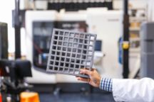 一名男子在实验室的3D打印机前拿着一个正方形交叉的3D打印灰色材料