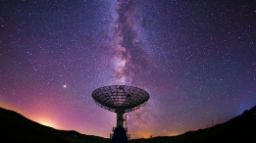 望远镜的筒形管映衬着繁星满天的夜空，银河呈现出紫色、蓝色、橙色和粉红色