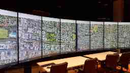 Inside a large room two long tables with four office chairs behind them are set up in front of a wide curving wall of 8 tall screens, all displaying a single image that is an aerial view of houses and streets.