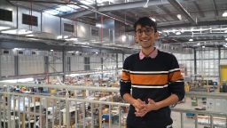照片中，一名身穿橙色横条纹黑色长衫的高个男子站在工厂环境中，背景是机器
