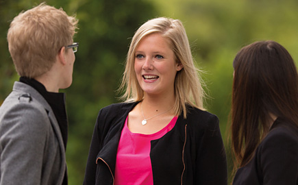 三个学生正在外面谈话。镜头聚焦于一个金发女郎夫人穿着一件黑色夹克和粉红色上衣。