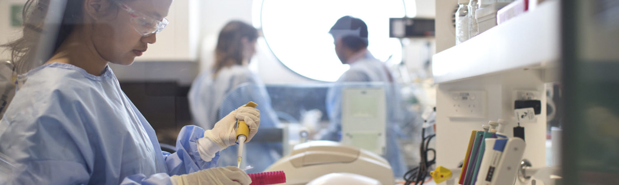 照片中，一个女孩戴着护目镜，穿着蓝色的手术服，在实验室里摆弄设备。背景中另外两名穿着工作服的学生的照片焦点不清。