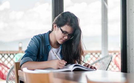 一位年轻女子手里拿着笔，正在看书。她坐在一张长木桌旁，身后有一扇窗户。